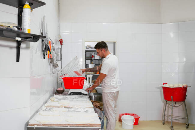 Side view of concentrated young male artisan in casual clothes and eyeglasses weighing ingredients on scales while standing at tale covered with flour in kitchen of modern bakery — Stock Photo