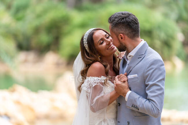 Seitenansicht des glücklichen jungen Ehepaares in Hochzeitskleidung stehend und Händchenhaltend in der Nähe des Sees und der grünen Bäume tagsüber im Garten — Stockfoto
