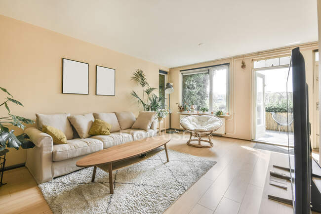 Interior de la luz sala de estar con ventanas francesas sillón de ratán sofá beige con cojines flores interiores y madera oval mesa TV y alfombra en el suelo - foto de stock