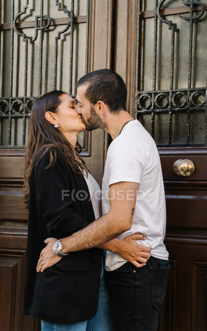 Вид сбоку на влюбленную молодую этническую пару в повседневной одежде, обнимающую друг друга и целующуюся, стоящую у дверей на городской улице — стоковое фото
