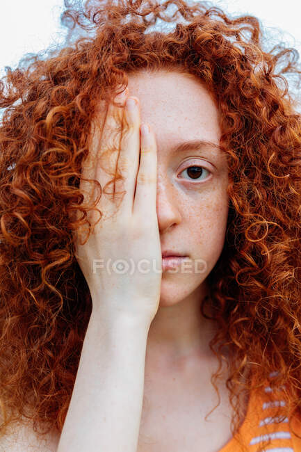 Jovem ruiva encaracolada fêmea com olhos castanhos e sardas cobrindo o olho com a mão e olhando para a câmera — Fotografia de Stock