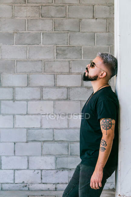 Side view of pensive male with beard and tattoos on arms in black outfit and sunglasses standing and leaning on grey wall of building in daylight — Stock Photo