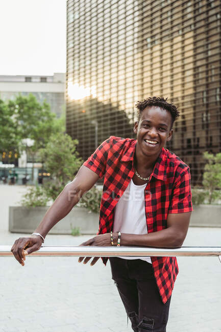 Happy African American male in checkered shirt leaning on metal railing and laughing brightly in urban park — Stock Photo