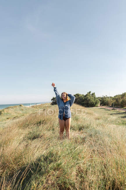 Preto e branco de mulher anônima de camisa de pé com braços levantados no prado gramado sob céu sem nuvens no verão olhando para a câmera — Fotografia de Stock