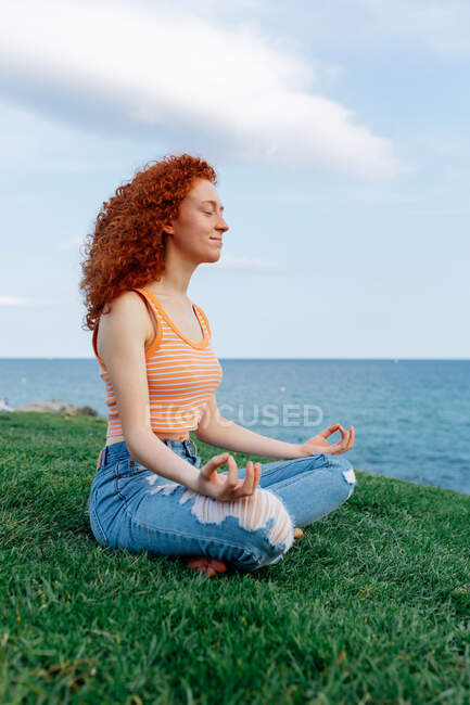 Side view of carefree female with long ginger hair in Padmasana pose with zen gesture on grassy hill on seashore — Stock Photo