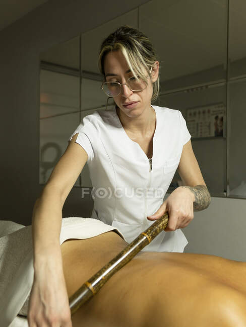 Female masseur using bamboo stick while massaging back of client in towel on couch in light salon — Stock Photo