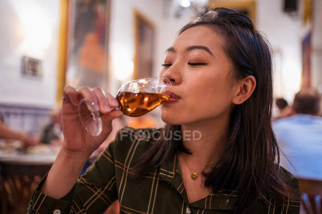 Atractiva hembra asiática con cabello negro bebiendo alcohol de la copa de vino durante la cena mientras está sentada en un restaurante con gente de fondo - foto de stock