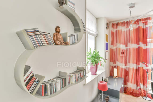 Moderna estantería blanca con colección de libros en la pared en la habitación elegante luz con planta verde y cortina de colores en el apartamento - foto de stock