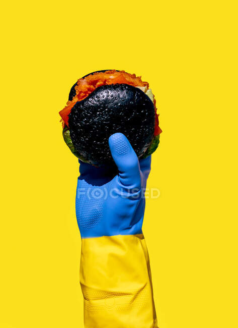 Erntehelfer im bunten Gummihandschuh demonstriert Burger mit Schwarzbrötchen als ungesundes Ernährungskonzept vor gelbem Hintergrund — Stockfoto