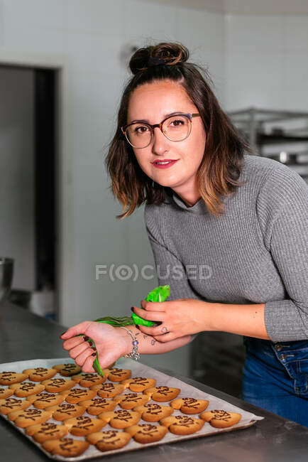 Вид збоку молода жінка-кондитерка з темним волоссям в повсякденному одязі, що прикрашає цукрове печиво у формі серця з шоколадною глазур'ю, що стоїть за столом у пекарні — стокове фото