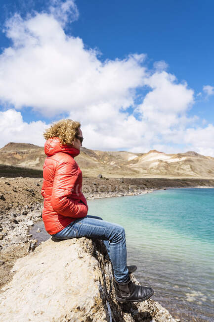 Side view of calm traveler sitting on edge of cliff above blue ocean against mountains in Iceland in sunny day — Stock Photo