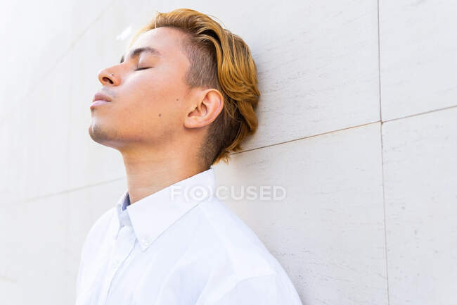 Grave giovane maschio in camicia bianca con gli occhi chiusi mentre si appoggia sul muro sulla strada in città — Foto stock