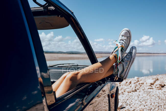 Crop feminino irreconhecível refrigeração no assento do passageiro no carro com as pernas na janela aberta na costa pedregosa da lagoa — Fotografia de Stock