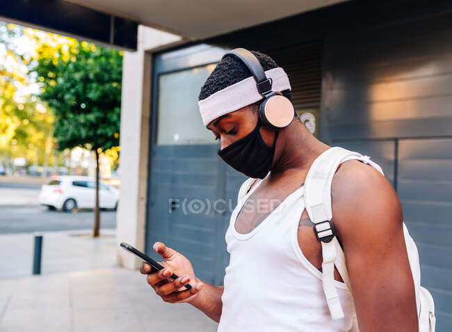 Vista lateral do homem afro-americano em máscara protetora e fones de ouvido em pé na rua e na Internet de surf no celular durante a pandemia de coronavírus — Fotografia de Stock