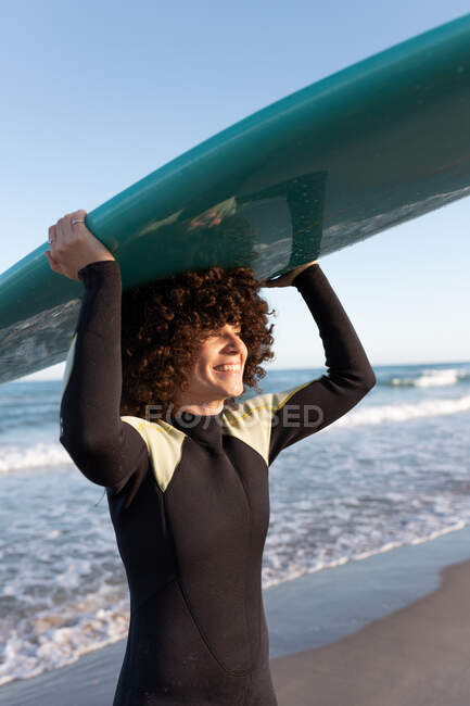 Vista laterale della giovane surfista felice in muta con tavola da surf in piedi che tiene la tavola da surf sopra la testa guardando lontano sulla riva del mare lavata dal mare ondulato — Foto stock