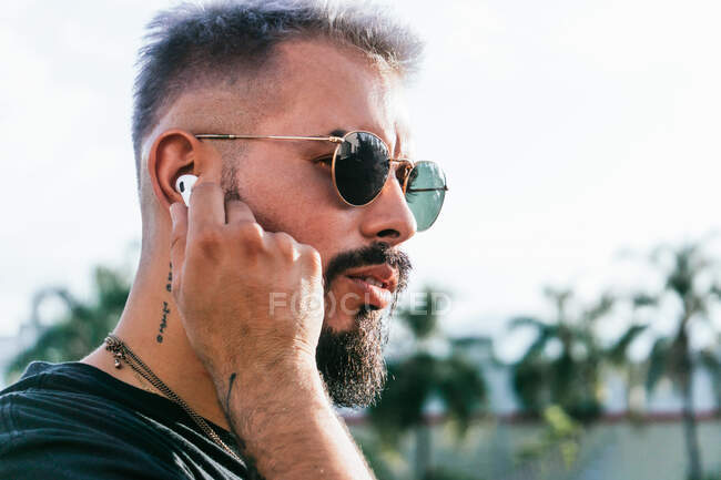 Side view of thoughtful bearded male with tattoos in casual clothes and sunglasses wearing earphones and looking away on street against green trees in sunny day — Stock Photo