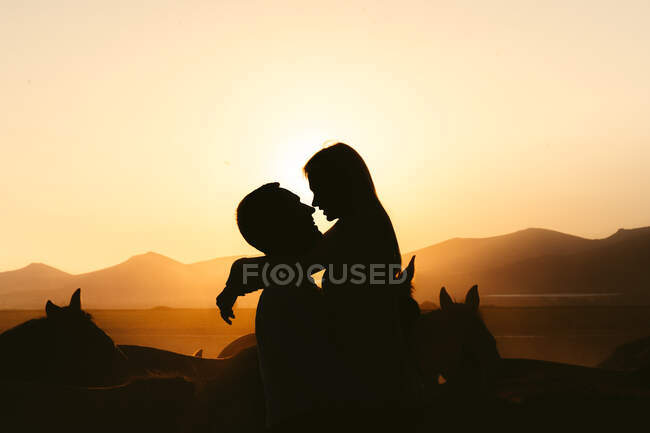 Vue latérale des silhouettes d'un couple amoureux s'embrassant tout en passant du temps ensemble sur un pâturage près des chevaux contre les montagnes au coucher du soleil — Photo de stock