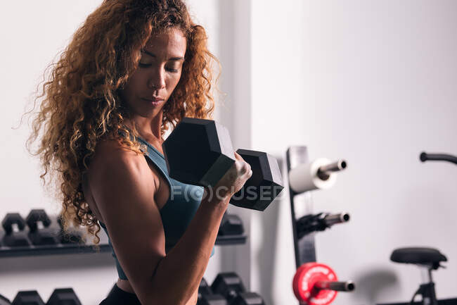 Vista lateral de esportista forte com cabelo encaracolado fazendo exercício no bíceps com haltere durante o treino no ginásio — Fotografia de Stock