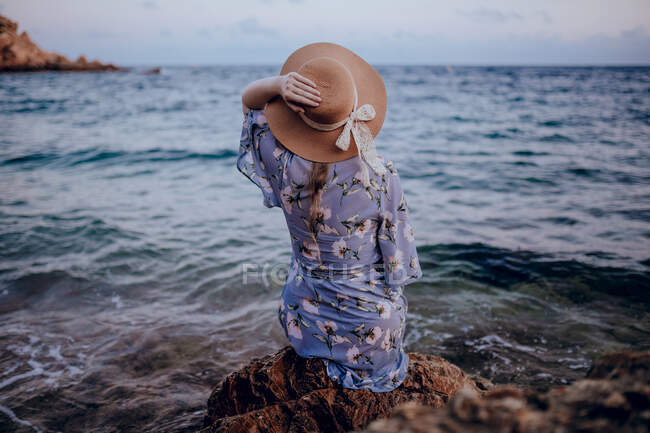 Visão traseira de uma jovem fêmea anônima em vestido de verão e chapéu sentado na costa rochosa enquanto olha para longe na noite de verão — Fotografia de Stock