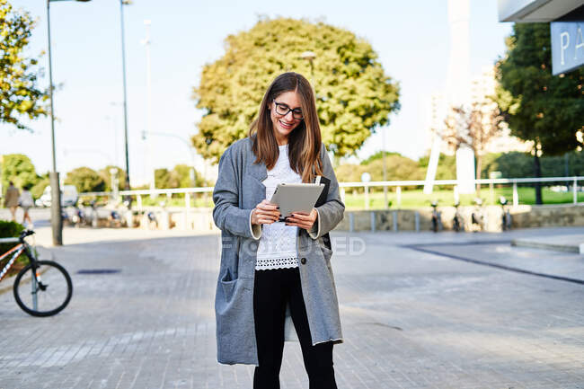 Happy female worker in eyeglasses surfing tablet while standing on street and doing job issues — Stock Photo