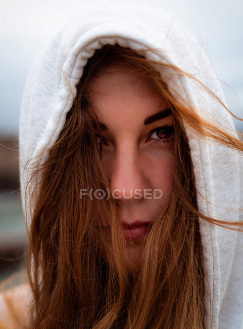 Portrait de charmante femme énigmatique aux cheveux roux volants et à la capuche regardant la caméra lors d'une journée venteuse en Galice — Photo de stock