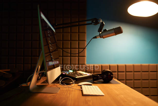 Computador moderno e teclado colocados em mesa de madeira com microfone em tripé e fones de ouvido em estúdio de gravação podcast escuro — Fotografia de Stock