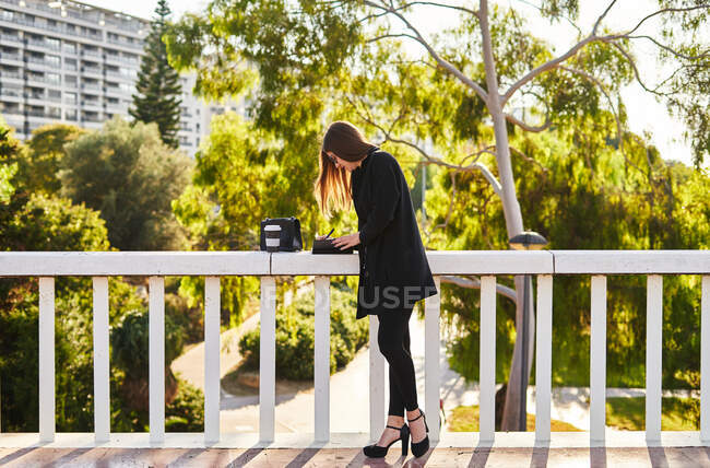 Concentrated businesswoman with brown hair in stylish clothes standing near concrete fence with cup of takeaway coffee and bag and whiting notes in notebook on street in sunny day — Stock Photo