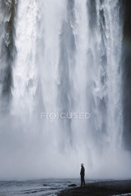 Vue latérale du randonneur masculin en vêtements décontractés debout au bord de la rivière sous la cascade spectaculaire Skogafoss coulant à travers une falaise rocheuse massive couverte de mousse verte — Photo de stock