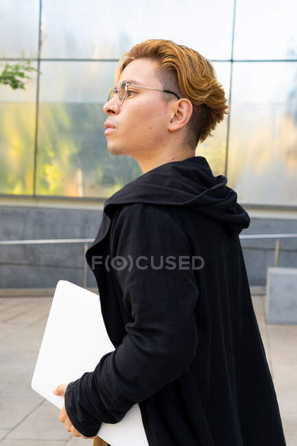 Вид сбоку студента в повседневной одежде и очках с современным нетбуком в руке, идущего по улице возле современного здания — стоковое фото