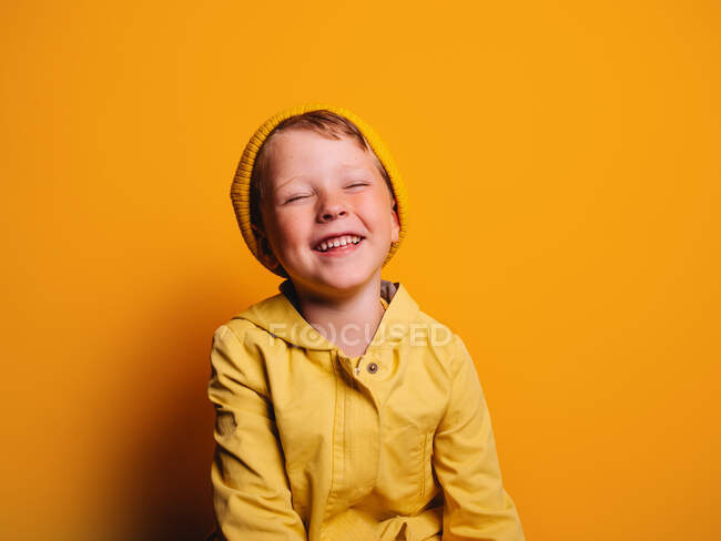 Happy boy in vivid yellow raincoat jacket and beanie hat laughing with eyes closed against yellow background in studio — Stock Photo