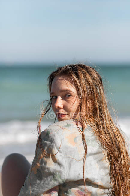 Mujer seria con el pelo mojado y ropa mojada de pie cerca del mar mientras mira a la cámara en el día soleado de verano - foto de stock
