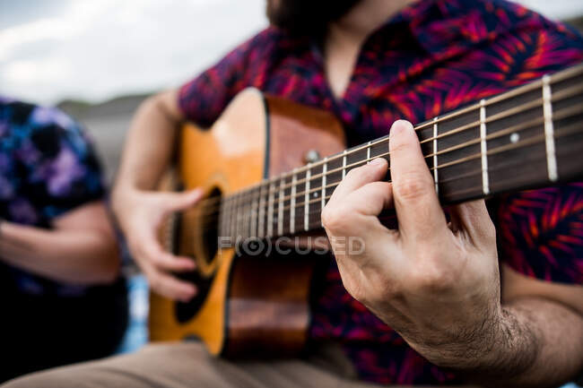 Неузнаваемый талант делает музыканта в повседневной одежде, играющего на акустической гитаре и поющего песни, сидя на песчаном пляже на природе при дневном свете — стоковое фото