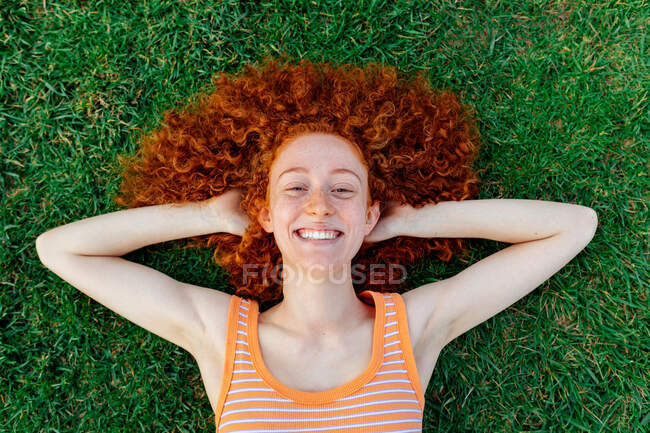 Вид зверху на щасливу кучеряву волохату жінку з руками позаду голови, що розслабляється на газоні і дивиться на камеру — стокове фото