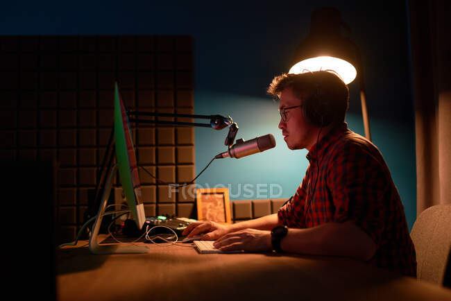 Side view of focused young male in checkered shirt and eyeglasses using computer and speaking in mic while recording podcast in dark studio — Stock Photo
