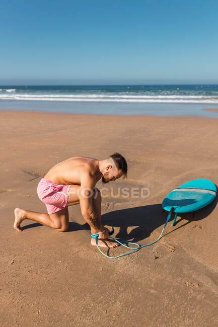Ganzkörper-Seitenansicht eines aktiven Mannes in Badehosen, der ein Beinseil anlegt, während er sich auf das Surfen am Sandstrand in Meeresnähe vorbereitet — Stockfoto