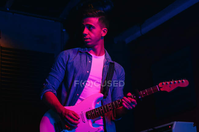Серьезный молодой человек, играющий на бас-гитаре во время выступления в легком клубе с неоновой подсветкой — стоковое фото