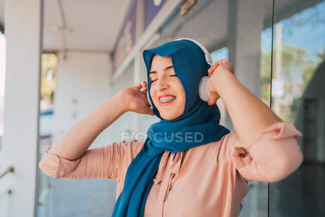 Femme arabe positive en foulard et écouteurs écouter de la musique avec les yeux fermés et profiter de chansons tout en se tenant dans la rue de la ville — Photo de stock