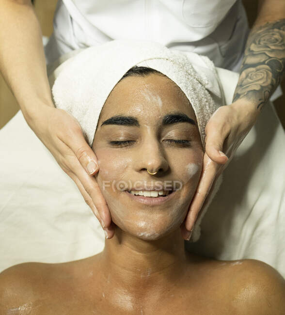 Masajista femenina aplicando crema en la cara del cliente mientras hace masaje facial en el sofá en el salón de spa brillante - foto de stock