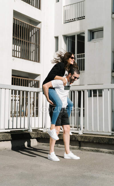 Corpo inteiro de feliz jovem etnia cara em roupas casuais e óculos de sol sorrindo ao dar passeio de piggyback para namorada amada perto de construção na rua — Fotografia de Stock