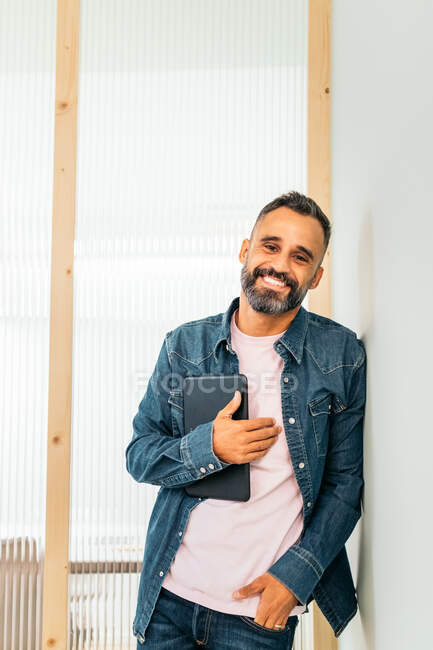 Empleado masculino hispano adulto barbudo seguro de sí mismo en traje casual sonriendo felizmente y mirando a la cámara mientras se apoya en la pared blanca con la tableta en la mano en el pasillo de la oficina moderna - foto de stock