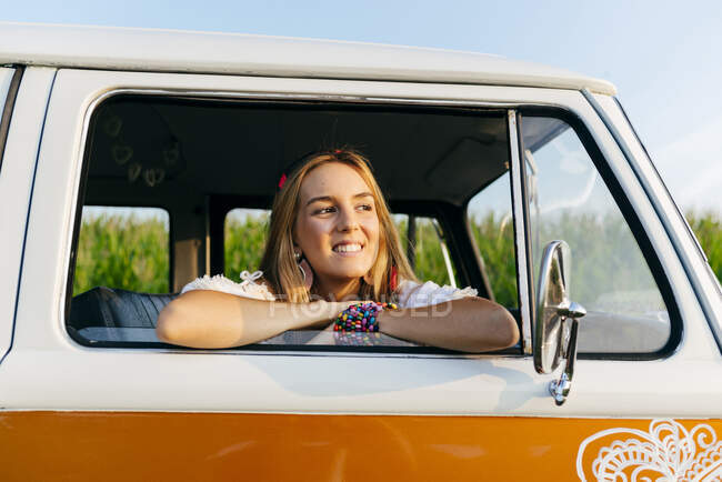 Menina loira bonito sentado dentro de uma van vintage e inclinando-se na janela em um dia ensolarado — Fotografia de Stock