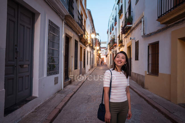 Viajera asiática positiva observando casas residenciales envejecidas mientras está parada en una calle estrecha durante un viaje en la ciudad de Córdoba en España - foto de stock
