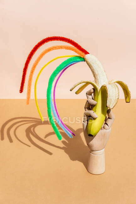 Mano maniquí de madera con salpicaduras de colores que salen de plátano pelado maduro colocado en la mesa contra fondo naranja en estudio de luz - foto de stock