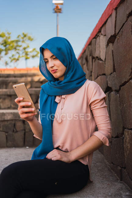 Content charming Arab female in traditional headscarf messaging on cellphone while sitting on stone bench in city park — Stock Photo