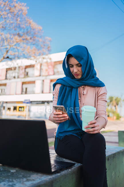 Contenu Arabe femme pigiste dans la navigation headfast téléphone mobile tout en étant assis sur le banc avec ordinateur portable et prendre une pause pendant le travail à distance — Photo de stock