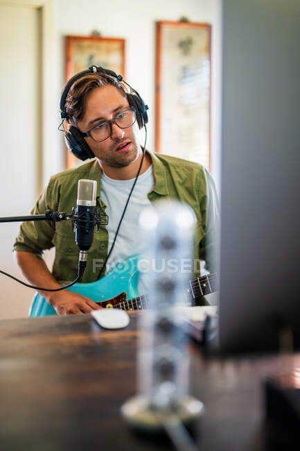 Männlicher Gitarrist mit Brille und Kopfhörer, der Instrument hält, während er Computer im modernen Studio benutzt — Stockfoto
