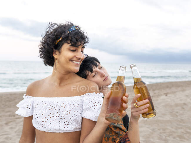 Amar casal multiétnico de fêmeas batendo garrafas de cerveja e desfrutar do dia de verão na costa — Fotografia de Stock