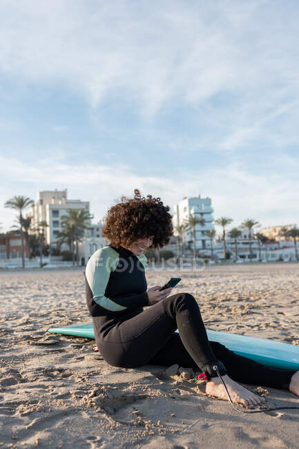 Mujer positiva con el pelo rizado usando el teléfono inteligente de navegación traje de neopreno mientras está sentado en la playa de arena con tabla de surf - foto de stock
