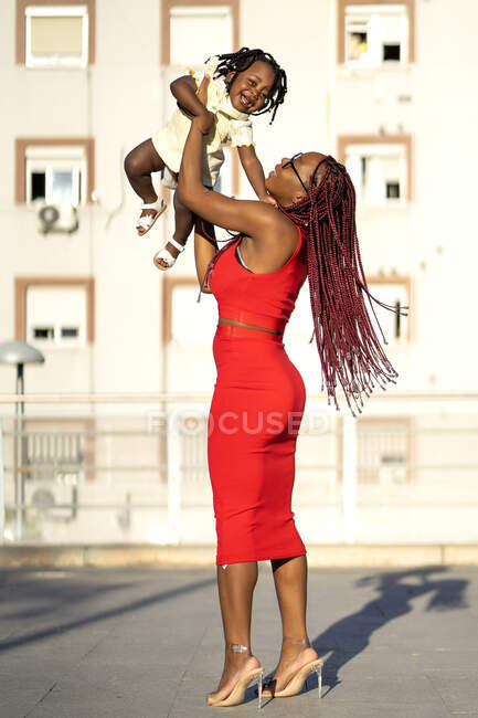 Vista laterale della femmina afroamericana con lunghe trecce in abito elegante in piedi e sollevamento felice figlioletta mentre si diverte in strada nella giornata di sole — Foto stock