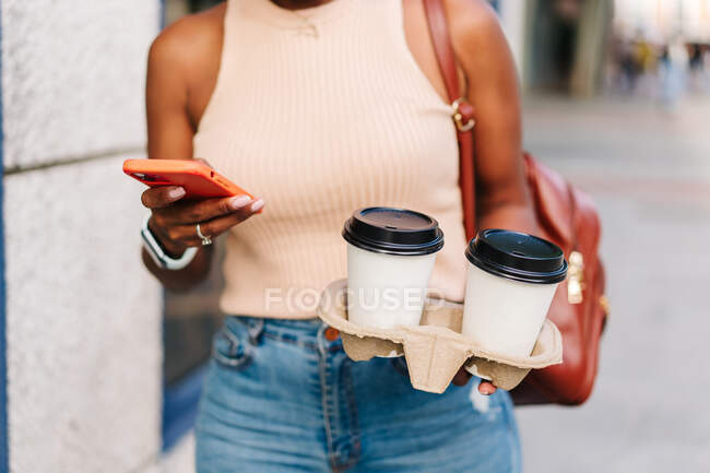 Anónimo de moda joven étnica milenaria en traje elegante y mochila con teléfono móvil mientras camina por la calle de la ciudad con tazas de café para llevar - foto de stock